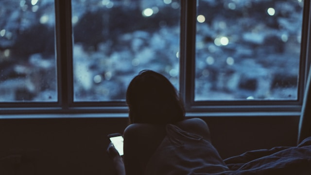 Woman sitting by a window at night looking at her phone